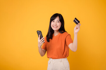 Confident Asian woman 30s in orange shirt, using smartphone and holding credit card on yellow...