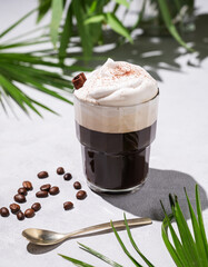 Iced coffee in a glass with foam on a light background with palm leaves, coffee beans and morning shadows.