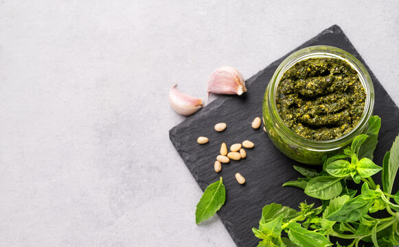 Traditional Italian pesto in a jar with green basil, pine nuts on a slate board on a light background. A classic sauce for spaghetti or bruschetta. Delicious vegetarian homemade food.