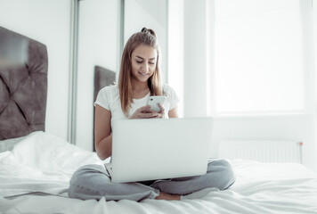 Young hipster woman using smartphone and laptop while sitting on bed in bright room. Modern gadgets