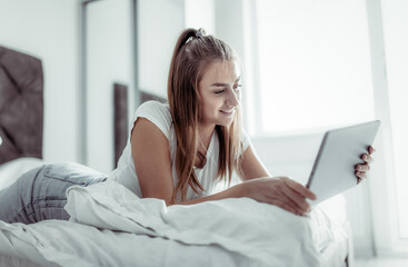 Young beautiful caucasian woman uses a tablet while lying on a bed in a bright room