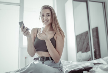 Smiling woman with lipstick and mirror sitting on bed in bright room
