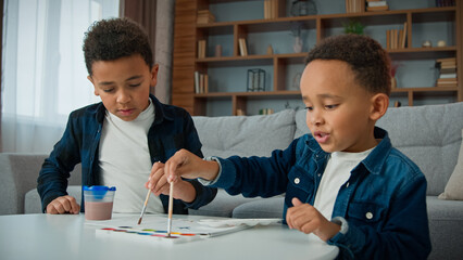 Two little children boys painting at home together. Ethnic brothers African American kids sibling paint picture with acrylic watercolor colors schoolboys indoors enjoy art hobby with paintbrushes