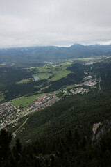 Fototapeta premium Aerial view of Mittenwald town surrounded by dense forest, green fields, and distant mountain ridge, captured from the Mittenwald High Trail under a cloudy sky.
