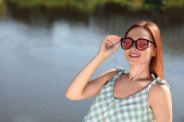 Beautiful woman in sunglasses near river, space for text