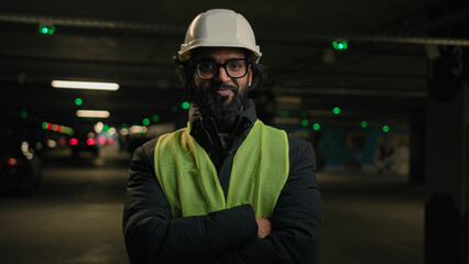 Indian man handyman worker contractor hands crossed posing in parking smiling mechanic in helmet, glasses power pose at factory warehouse manufacture. Arabian male builder engineer fitter in hard hat