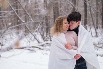 A young couple walks in the park in winter. The guy and the girl are kissing wrapped in a white blanket outdoors.