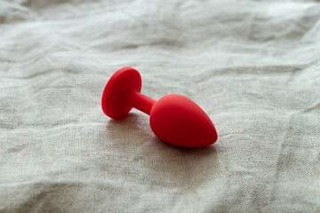 Red silicone anal plug, sex toy, on a beige bed sheet in the bedroom. Contrast photo of anal toys, erotic toys - obrazy, fototapety, plakaty