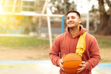 Young man with basketball smiling on playing court with sweatshirt and t-shirt on his shoulder. Winning team concept.