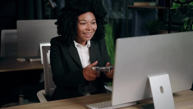 Excited young african american businesswoman gamer sitting at desk on workplace and playing in video games on computer indoors Happy curly office worker controlling joystick at late evening alone