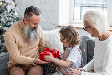 Attractive senior grandparents exchanging Christmas and New Year gift box with granddaughter, cute little girl. Looking happy and excited sitting near decorated tree in red Santa hats. Family time