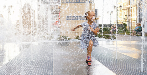 Banner Child playing in water fountain in the city. Hot summer weather. Happy girl playing with water sprinklers, running and laughing. Active childhood. Games outdoors. Modern city street - obrazy, fototapety, plakaty