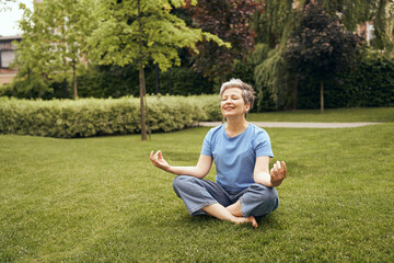 Senior woman in lotus pose sitting on green grass while doing yoga at city park. Healthy lifestyle