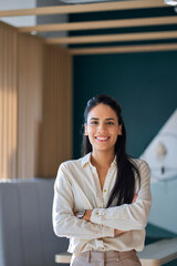 Confident happy professional young latin business woman company employee, lady executive manager, smiling female worker or entrepreneur looking at camera standing in modern office, vertical portrait.