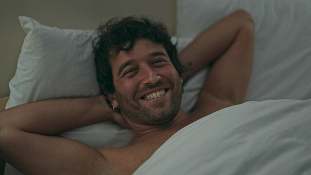 Young hispanic man relaxed on bed smiling shirtless at bedroom