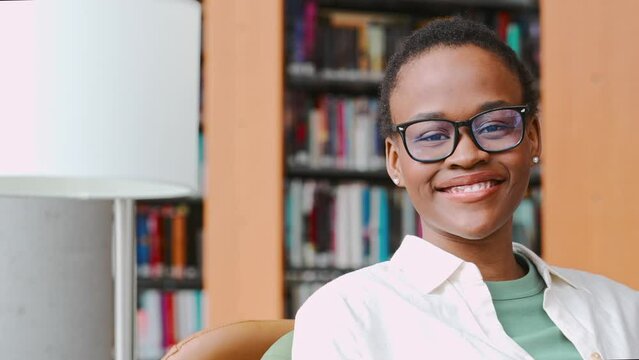 Happy cute African teen girl, smiling short-haired cute Black ethnic college student wearing eyeglasses looking at camera in modern university campus library. Close up face portrait.