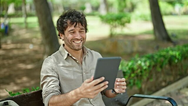 Young hispanic man make selfie by touchpad sitting on bench smiling at park