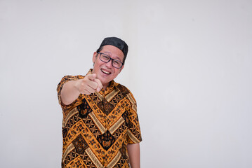 A southeast asian man mocking and ribbing someone. Wearing a batik shirt and songkok skull cap with hand on chest. A middle aged muslim male in his 40s. Isolated on a white background.