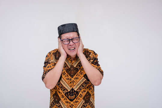 A southeast asian muslim man covering his ears from blaring noisy music. Wearing a batik shirt and songkok skull cap with hand on chest. Isolated on a white background.
