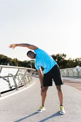Fotobehang Muscular african american man wearing blue t shirt warming up lening to the side before running © Maria Vitkovska