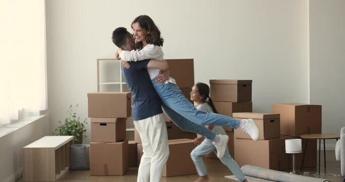 Cheerful dad lifting and spinning happy mom over floor at joyful active running kid girl, smiling, laughing, having fun, celebrating moving into new apartment among stacked cardboard boxes