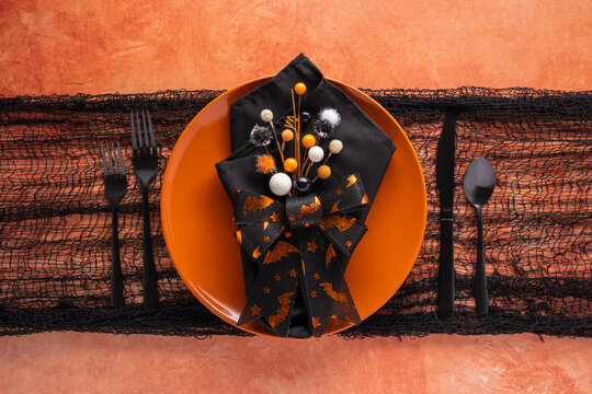 Halloween party table setting in black and orange with plate, naokin, flatware and decor on orange mottled background
