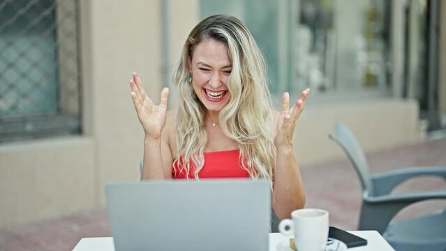 Young blonde woman using laptop sitting on table with winner gesture at coffee shop terrace