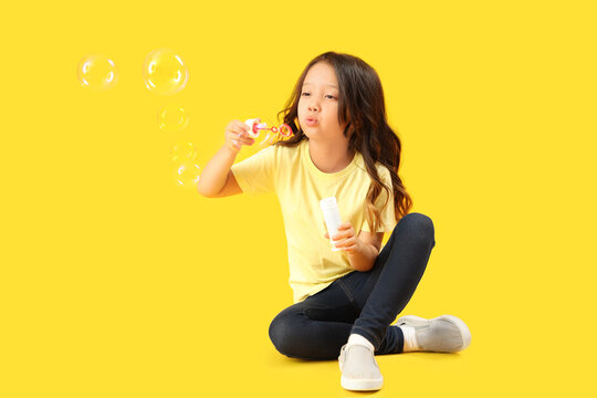 Cute little girl blowing soap bubbles on yellow background