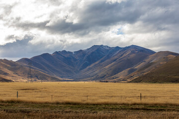 Fototapeta premium Photograph of a dry mountain range running behind a large brown agricultural field with low level grey clouds on the South Island of New Zealand