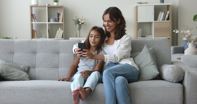 Positive young mother and pretty preschool girl using online media application on smartphone, taking funny selfie on mobile phone, self video for social network, watching content on Internet