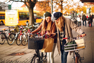 Young lesbian couple using a smart phone while shopping and pushing their bicycles on a city...
