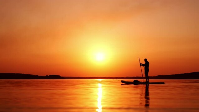 Orange sunset over the river. Silhouette of a male standing on the sup board and slowly rowing with a paddle.