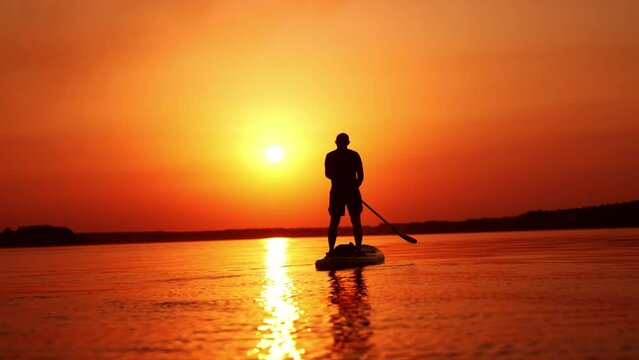 Man stands on the sup board facing the sunset. Man holds a paddle and row with it from each side.