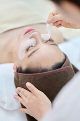 아시아 한국의 젊은 여성이 스파 또는 피부관리전문점 또는 피부과 병원에서 갈색 두건을 쓰고 침대에 누워 베이지색 대형 타올을 덮고 하얀색 셔츠를 입은 전문관리사가 눈팩으로 눈밑 다크서클과 주름관리를 하고있다. 