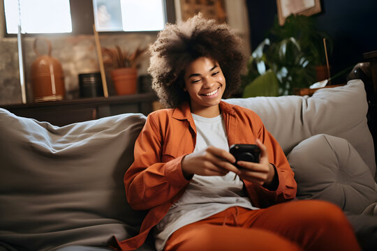 Happy relaxed gen z African American teen sitting on couch at home holding smartphone