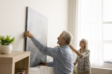 Cheerful senior couple decorating apartment, hanging large picture on white wall, laughing, having...