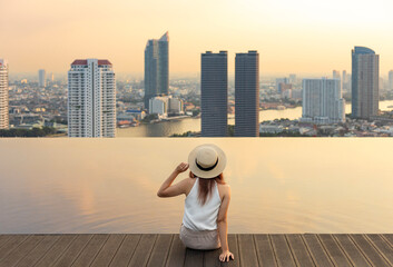 Pretty tourist woman is relaxing and enjoying her rooftop swimming pool view with cityscape background for vacation and travel at the sunset with skyscraper and downtown highrise condominium around - obrazy, fototapety, plakaty