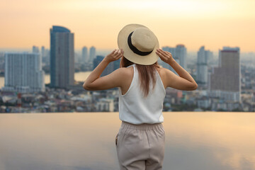 Pretty tourist woman is relaxing and enjoying her rooftop swimming pool view with cityscape...