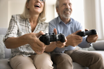 Hands of joyful elderly husband and wife pushing buttons on gamepad joysticks, playing virtual fight, battle, online video game, sitting on sofa with joypads, laughing
