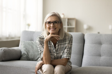 Happy attractive blonde mature woman in stylish eyeglasses sitting on couch, looking at camera,...