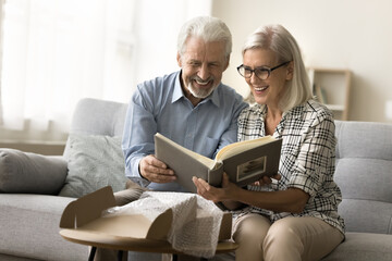 Cheerful elderly couple in love opening parcel, cardboard box at home, producing picture book,...