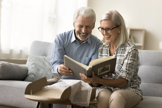 Cheerful elderly couple in love opening parcel, cardboard box at home, producing picture book, watching photographs in received family photo album with joy, smiling, enjoying memories, nostalgia