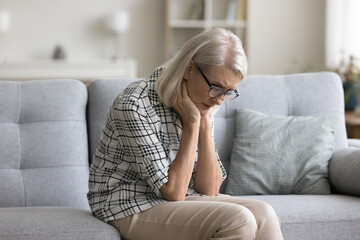 Sad depressed retired blonde woman sitting on sofa at home, looking down in deep bad thoughts, leaning chin on hands, thinking on healthcare problems, going through grief, loss, loneliness