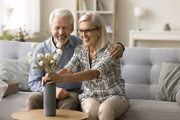 Happy senior wife getting bunch of flowers from loving husband due to birthday, putting bouquet into vase. Positive older retired couple celebrating anniversary, special day