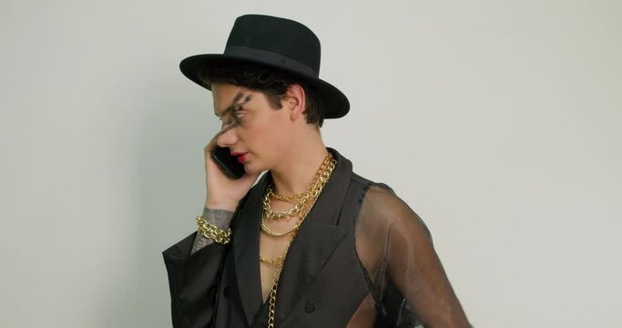 transgender man is in makeup talking on cell smart phone gray background Communication Gorgeous american gay in black fashionable outfit, chain and hat accessories having conversation lgbt lifestyle