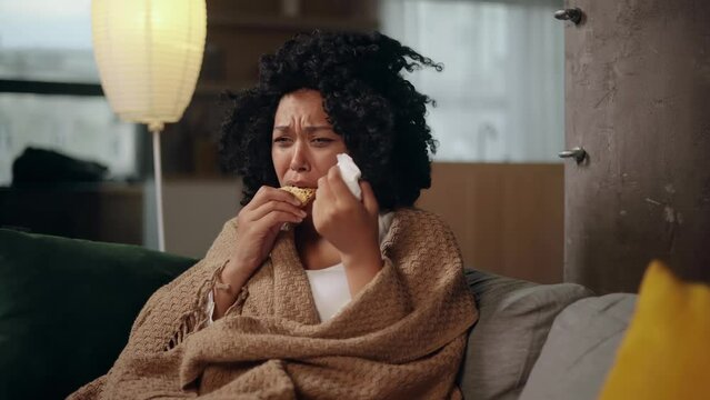Portrait of sad young woman dealing with stress by eating food sitting on sofa at home Upset female wrapped in blanket crying and eating cookies indoors alone Mental heath problem