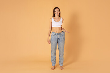 Diet, slimming concept. Happy young european lady wearing big jeans after weight loss diet program, posing on beige background, full length copy space © Home-stock