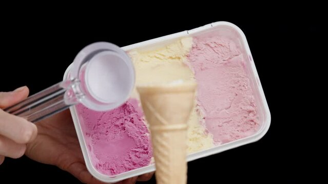 A container of ice cream in pink tones and a spoon to scoop it into the waffle cone, isolated on a black background.