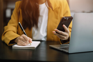 Female businessman use a smartphone and take notes in the notebook at the office desk in the morning.