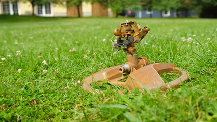 Old rusty metal lawn sprinkler on green grass. Houses in the background. Selective focus.